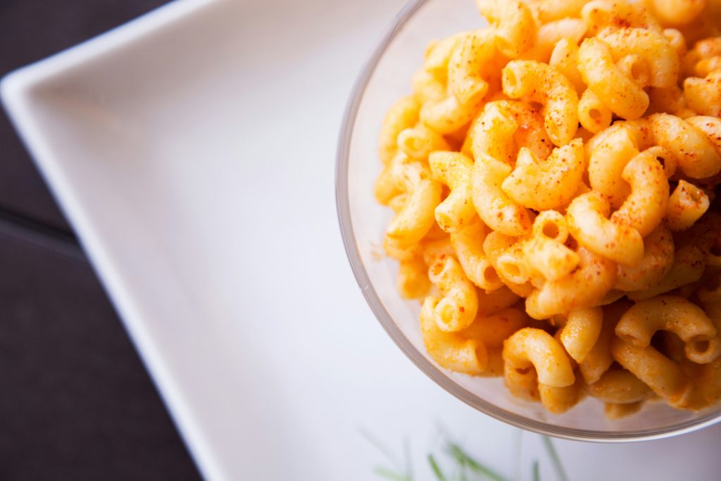 A clear glass bowl full of cheesy elbow pasta, dusted with paprika, on top of a square white plate.